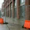 Stalen bouwschutting op een Nederlandse stoep, geflankeerd door oranje-witte veiligheidsbarricades en een bakstenen gevel op de achtergrond.