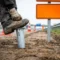 Gegalvaniseerde grondanker in wegbermgrond getrapt door werkhandschoen, oranje tijdelijk verkeersbord en verkeerskegels op Nederlandse snelweg op achtergrond.