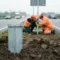 Verzinkte stalen grondpot ingegraven langs Belgische autoweg, arbeiders in veiligheidsvesten plaatsen verkeersbord bij bewolkt daglicht.