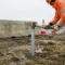 Bouwvakker in hesje slaat stalen grondpot in Europese bouwgrond, betonnen barrier en oranje verkeerskegels op achtergrond.