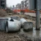 Verzwaarde gegalvaniseerde grondpotten op bouwplaats, één ingestort in vers beton naast een stalen verkeersbordpaal, grindbodem op de achtergrond.
