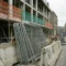 Gestapelde stalen bouwschuttingen tegen een betonnen barrier op een Europese stedelijke bouwplaats, met steigers en veiligheidsnet op de achtergrond.
